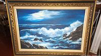 Full view of the framed oil painting showing seascape with crashing waves, rocky coastline and partly cloudy sky in hues of blue and white.