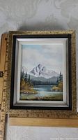 Framed landscape painting with a mountain, pine trees, lake, under cloudy sky. Rulers placed to show frame size of about 8 x 6 inches.