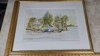 Framed watercolor artwork showing cottage, trees, bridge, and boat by lake with ruler for size reference.
