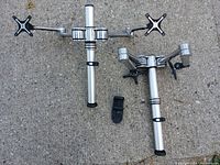 Two adjustable dual-arm metal computer monitor stands laid on concrete surface, showing VESA mount brackets and base clamps.