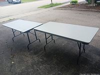 Two folding tables set outdoors on pavement. Light gray tabletops with black folding legs visible. Both tables appear sturdy and in good condition.