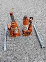 Two orange Strongarm hydraulic jacks side by side on concrete, each with metal pump handles beside them.