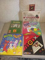 Photo showing six assorted board games stacked on a couch including Mucca Pazza, Mod X, Pamplona, Torres, Leg Los!, and Jernhest.