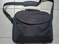 Black Dell laptop bag lying flat on tiled floor showing front side with flap and Dell logo in blue