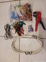 Photo showing assorted electrical materials including wire connectors in a plastic bag, multicolored alligator clip test leads, thin speaker wires, coaxial cable, and a red-handled wire stripping tool on a tiled floor.