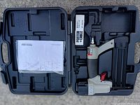 Top view of the open black plastic hard case containing the nailer, instruction manual, and an empty compartment for staples or accessories.