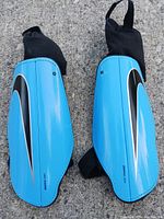 Pair of blue Nike shin guards placed on concrete floor, showing front curved hard outer shell with Nike swoosh and elastic straps.
