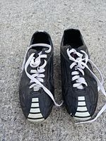 Top view of pair of black soccer cleats with white laces and white striping on toe area sitting on concrete surface