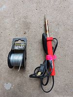 Soldering iron with red handle and solder wire spool shown on concrete ground, front angle.