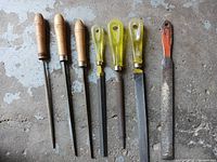 Overview of eight assorted metal files laid out horizontally on a concrete surface, showing wood, yellow plastic, and one orange-marked handles.