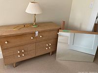 Wooden dresser with 6 drawers and a brass lamp on top, rectangular mirror placed beside dresser on floor