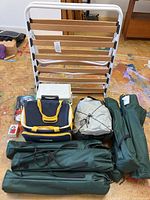 Fold out cot upright with wooden slats, cooler bag, cooler and three green camping chair bags on a floor