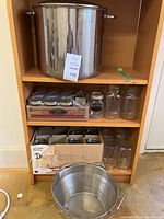 Wide view of shelving unit holding a large stainless steel canning pot on top shelf, various canning jars (quart and pint sizes) arranged on middle and lower shelves, and boxes containing lids and bands beneath jars. The floor is concrete; contents appear used but intact.