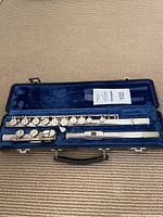 Open flute case showing three-piece silver-colored flute inside on a blue padded lining. Flute keys and lip plate visible. Case silver latches and black handle visible.