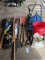Wide view of garden tools, netting rolls, tarp, spreader, rakes, shovels, watering can, gloves, and fertilizer bag arranged on floor in garage.