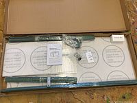 Opened box showing greenhouse metal frame parts, polycarbonate panels wrapped in protective film, instruction booklet, and hardware in plastic bags.