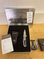 Branded metal tin open to show all contents: knife, nylon sheath, adapter kit, leather bit pouch and user guide