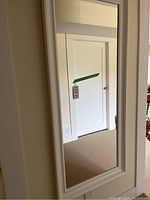 Front view of large white framed wall mirror with reflection of a white door and beige carpeted floor.