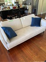 Fabric beige sofa with two blue cushions in a room with hardwood floor and kitchen in background