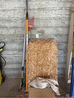 Full view showing straw bale, pole pruner with orange handle, apple picker with wire basket, branch cutter.