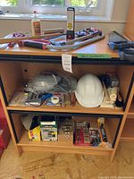 Full view of assortment on cabinet shelves showing saws, stapler, white hard hat, face shield, batteries, garbage bags, and other items
