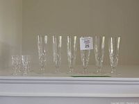 Overview: six tall crystal flutes arranged with two small cordial glasses at left on white shelf.