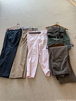 Photo of assorted women's trousers in various colors including black, beige, pink, gray, brown corduroy, and a green jacket along with a blue top on hangers.