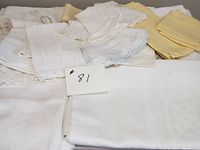 Overview of assortment of folded linens including tablecloths and napkins in mostly white and some yellow fabric