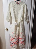 Front view of cream-colored quilted duster with floral print on lower half, zipper front, and sash tied at waist.