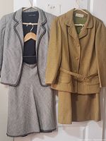Two vintage women's skirt suits hung side by side. Left suit is grey with black trim, collarless jacket and skirt. Right suit is tan with diamond texture, belted jacket and skirt.