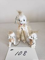 Front and partial side view of white ceramic dog figurine with two puppies attached by a gold metal chain. Each has tan fur accents on head, back and tail. Face details hand-painted. Displayed on white surface with lot number card.