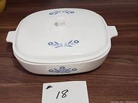 Front view of white Corningware Pyroceram covered casserole featuring blue floral designs on lid and sides, positioned on wood surface with label showing number 18.