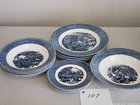 Blue and white Currier & Ives dinnerware pieces arranged showing four dinner plates, soup/cereal bowls, and side plates with scenic rural prints.