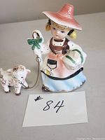 Side front view of ceramic figurine of girl Mary with basket and pink bonnet hat, linked by chain to lamb figurine.
