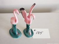 Front and side view of two pink flamingo ceramic figurines on green bases, one with wings down and one with wings raised.
