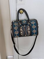 Full view of large rectangular handbag hanging on a door handle. Shows detailed embroidery in blue, white, and black with straps and charm.