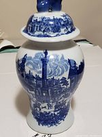 Front view of the tall ceramic jar with lid showing blue and white scenic architectural and figure patterns.