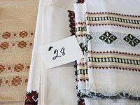 Close-up photo showing a stack of three different embroidered linen tablecloths with floral and geometric patterns, with a paper numbered 28 on top.