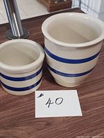 Two cream-colored Robinson Ransbottom Ohio pottery crocks with two blue stripes, one large and one small, displayed together.