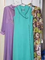 Three 1970s vintage maxi dresses hanging on hangers: lavender long-sleeve dress with waist embellishments, turquoise sleeveless dress with decorative buttons and trim, and a brown floral print long-sleeve dress with tags.