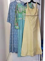 Photo of three vintage dresses hanging: blue paisley long-sleeve, green floral sheer top aqua bottom, and yellow sleeveless gown
