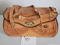 Front and side view of vintage tooled leather handbag showing the floral embossed design, brass clasp, and black whipstitched edges.