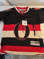 Front view of a youth small Ottawa Senators jersey in black, red, and white, showing large central O logo, patches on sleeves, and multiple autographs on the front.