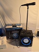 Front and side view of the Bluetooth karaoke speaker with LED illuminated speaker cone, control panel, and microphone stand setup with box in background.