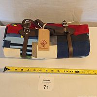 Folded throw blanket with visible leather carrying straps and Canadiana tag, placed next to measuring tape showing about 14 inches in width.