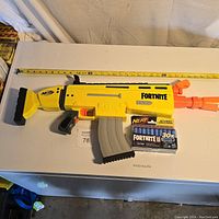 Full view of the yellow and orange Fortnite Nerf blaster next to the 30-pack of Fortnite Elite darts on a white surface