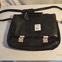 Front view of black leather laptop bag with two silver metal clasp closures and adjustable shoulder strap.