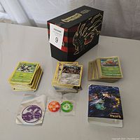 Photo of lots of Pokemon cards stacked in three piles with a decorative box standing behind. Also shows two round game tokens and a card sleeve with Pokemon design.