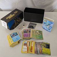 Overview photo showing the Pokemon-themed black storage box, multiple stacks of Pokemon trading cards, a card binder, and game coins.