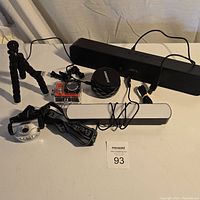 Overview of all electronic items in the lot on a table, showing the headlamp, action camera with waterproof case, two soundbars, flexible tripod, and an unknown round black item.